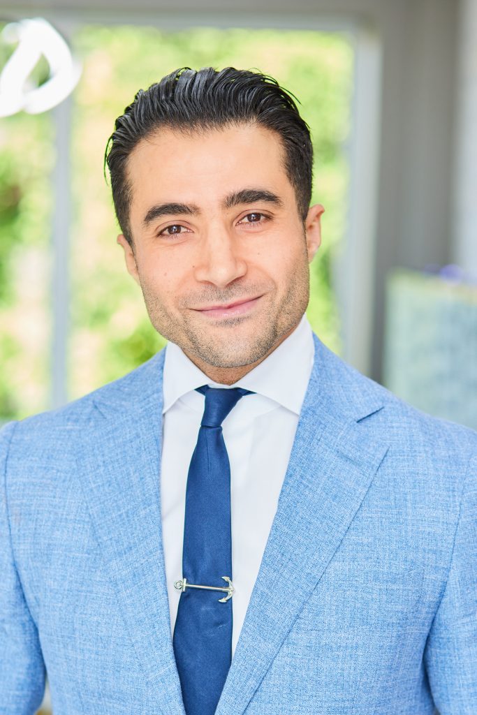 Dr. Faisel Ashtewi, prosthodontist, smiling in a blue suit and tie, in a modern dental practice setting, emphasizing expertise in dental implants and cosmetic dentistry.